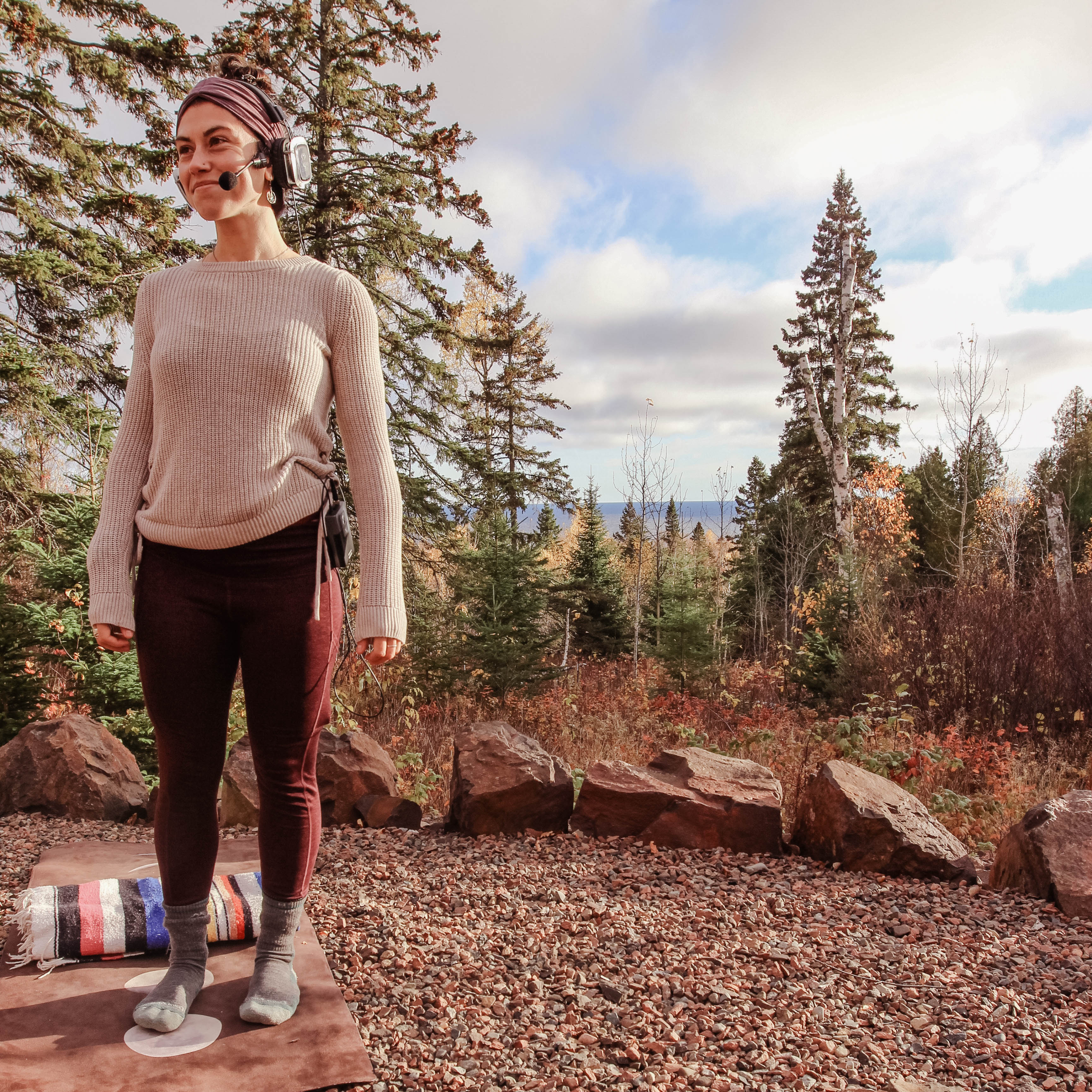 A woman standing outdoors on a rocky ground during daytime, with tall trees in the background and a partly cloudy sky. She is wearing a beige long-sleeve top, burgundy pants, gray socks, and a headset with a microphone, and is smiling.
