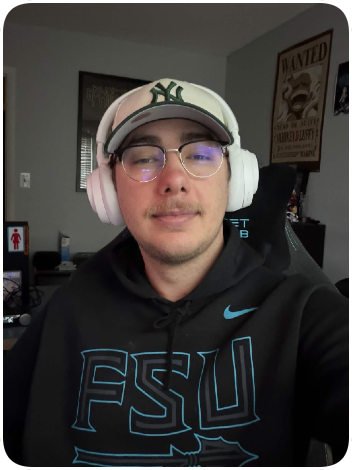 Young man wearing glasses, a baseball cap, and headphones, taking a selfie indoors with a black gaming chair and framed posters on the wall.