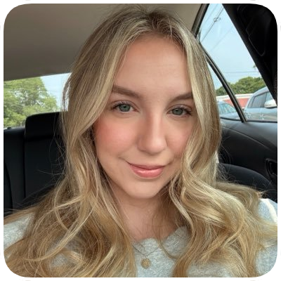 A young woman with blonde wavy hair taking a selfie inside a car, with trees visible through the window in the background.
