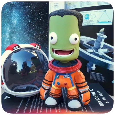 A plush toy of a green cartoon alien with big eyes and a wide smile, dressed in an orange spacesuit, standing next to a toy spacecraft with a reflective sphere and space-themed books in the background.