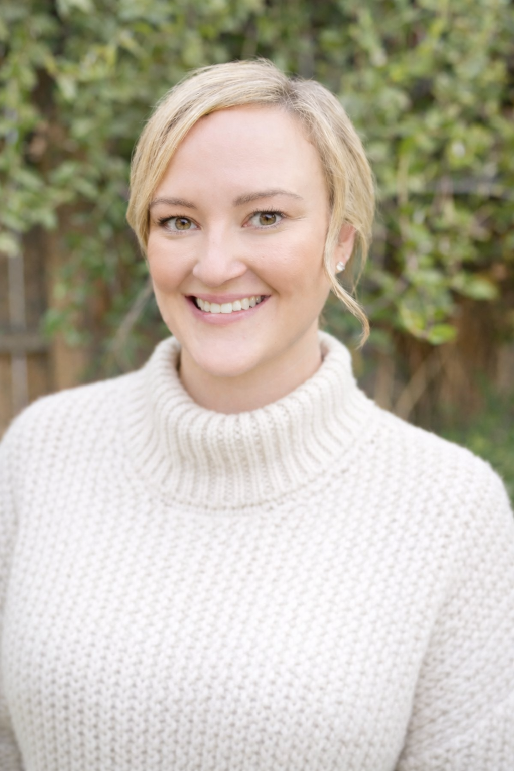 A woman with short blonde hair smiling outdoors, wearing a cream-colored knitted turtleneck sweater, with green foliage in the background.