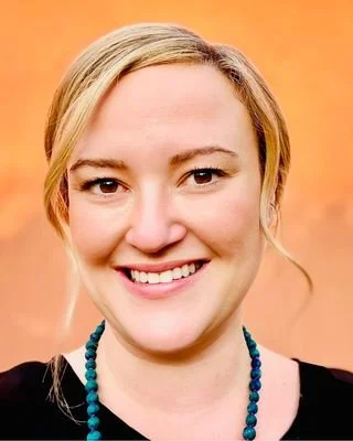 Close-up of a smiling woman with blonde hair and a beaded necklace against an orange background.