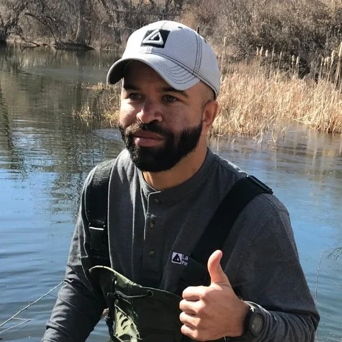 A man with a beard wearing a gray baseball cap and a dark gray long sleeve shirt giving a thumbs-up while fishing by a river with tall brown grass in the background.