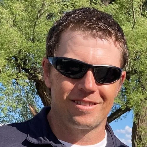 Charles Bryant wearing black sunglasses, smiling outside with trees and blue sky in the background.