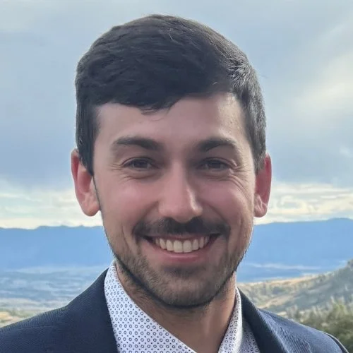 Liam Cullinane wearing a dark suit jacket and a white patterned shirt, standing outdoors with hills and a cloudy sky in the background.
