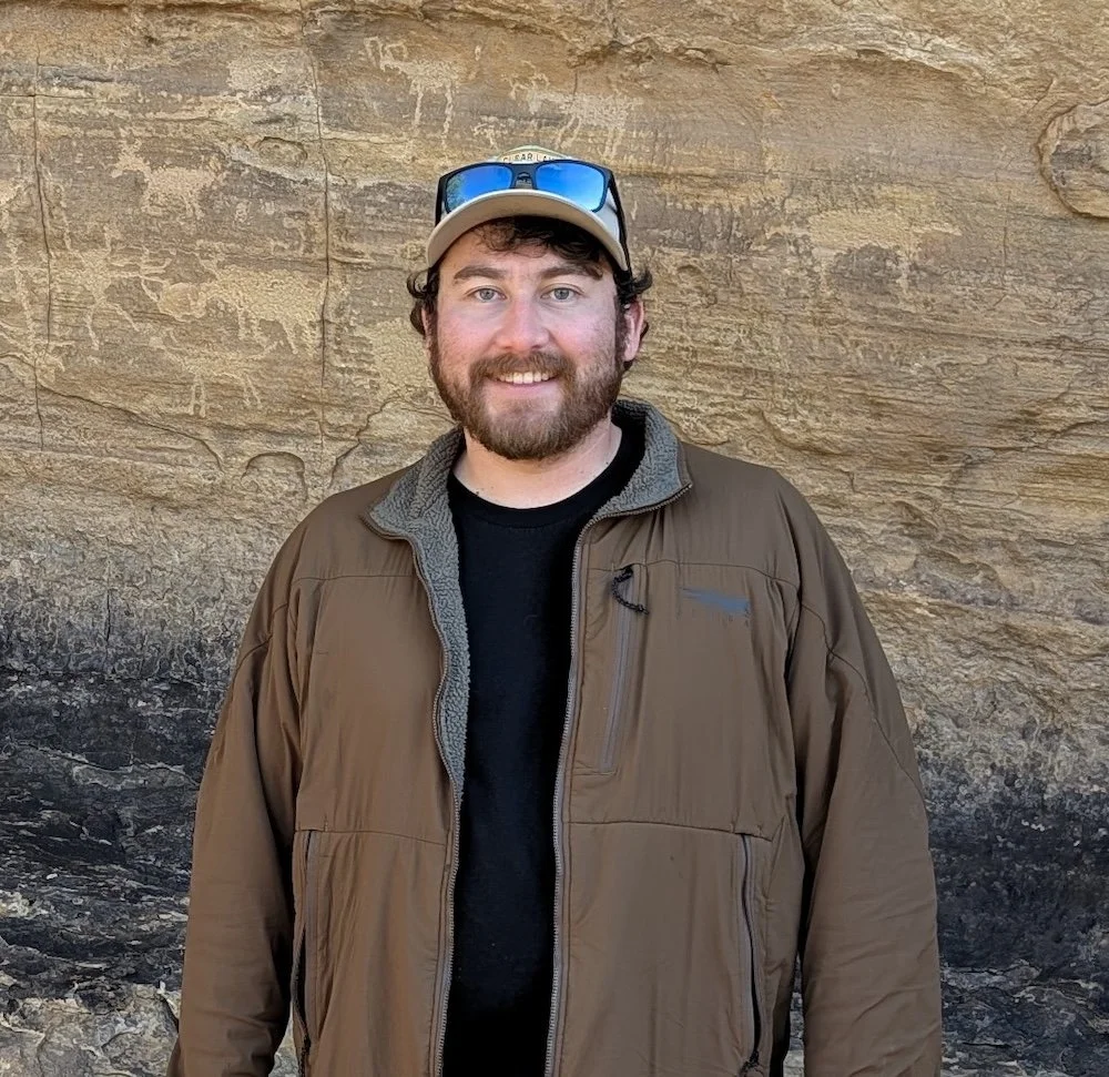 Nick Race standing in front of a rocky background, wearing a brown jacket, a black shirt, a beige cap, and sunglasses resting on the cap.