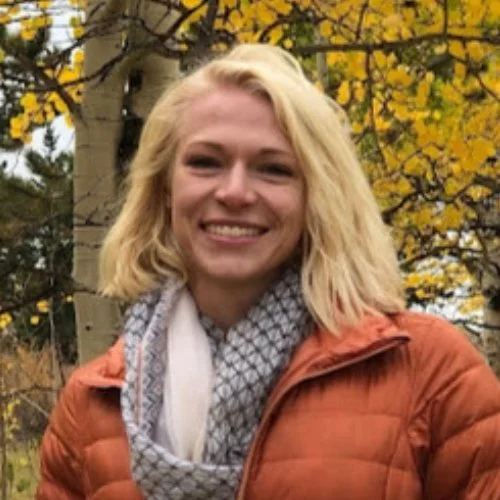 Lexie Sorensan smiling outdoors in front of a tree with yellow leaves, wearing an orange jacket and a gray patterned scarf.