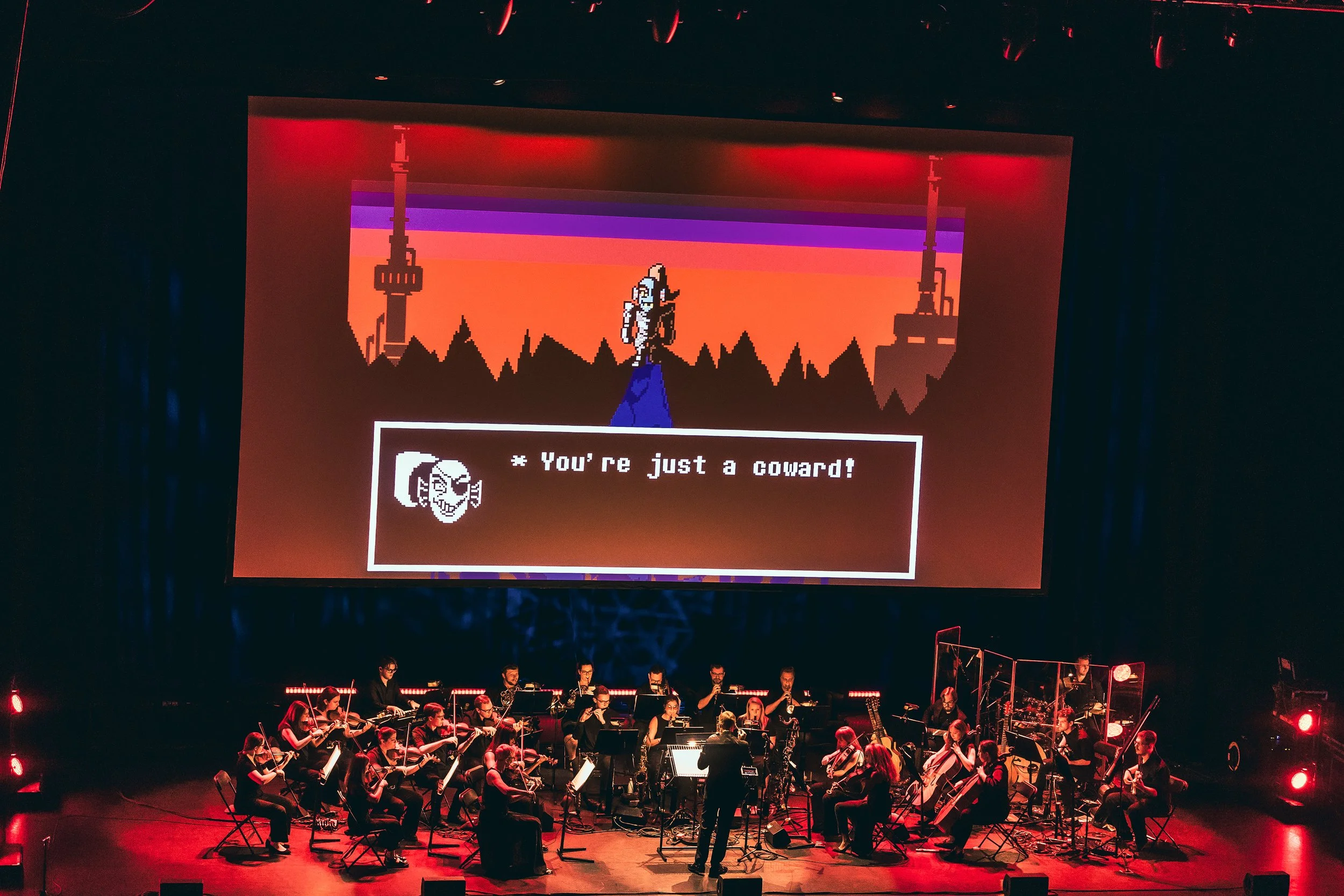 An orchestra performing on stage with a large screen projecting a pixel art scene from a video game. The scene shows a character with a speech box saying, "You're just a coward!" in front of a sunset skyline with towers.
