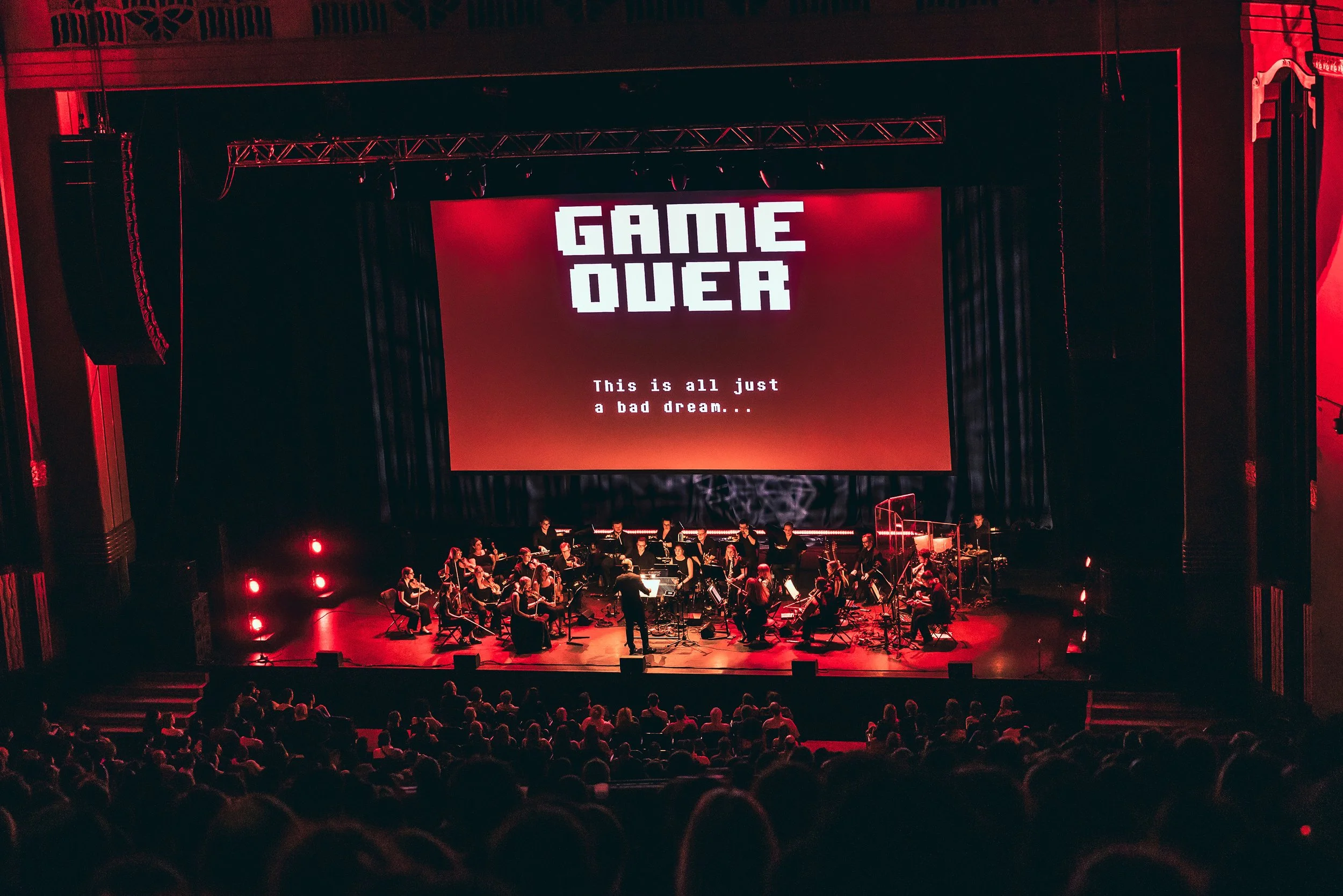 A stage with an orchestra performing, a large screen displays 'GAME OVER' and the message 'This is all just a bad dream...' in a dark theater setting with red lighting and an audience watching.