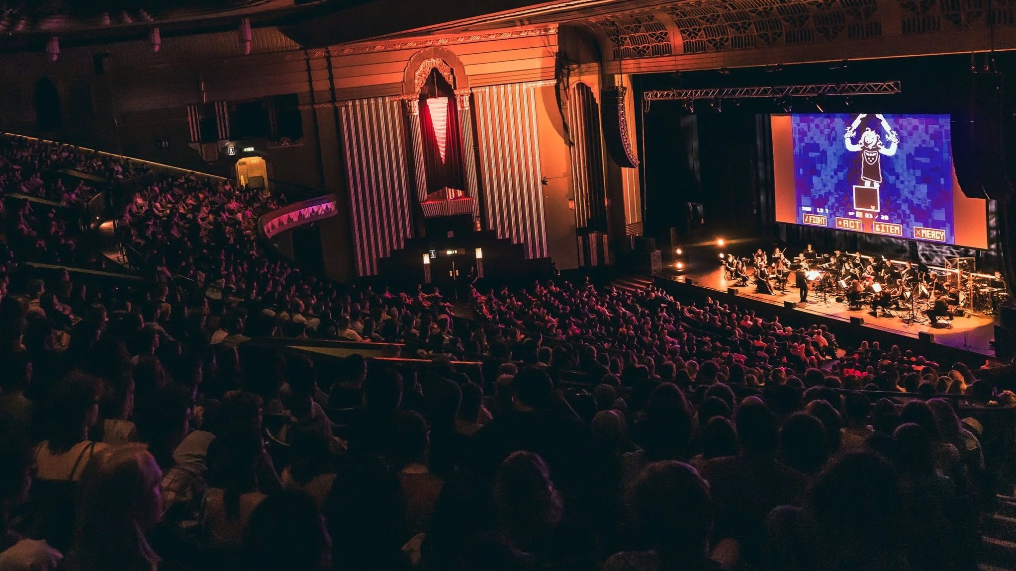 A crowded theater with a live performance on stage, featuring a large screen displaying a pixel art character and video game interface with options for Fight, Act, Item, and Mercy.