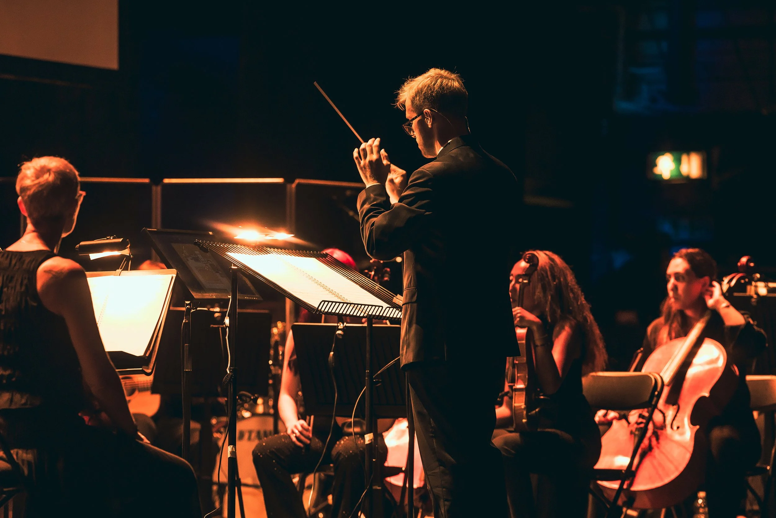 A conductor leading an orchestra during a performance, with musicians playing string and wind instruments in a dimly lit stage setting.