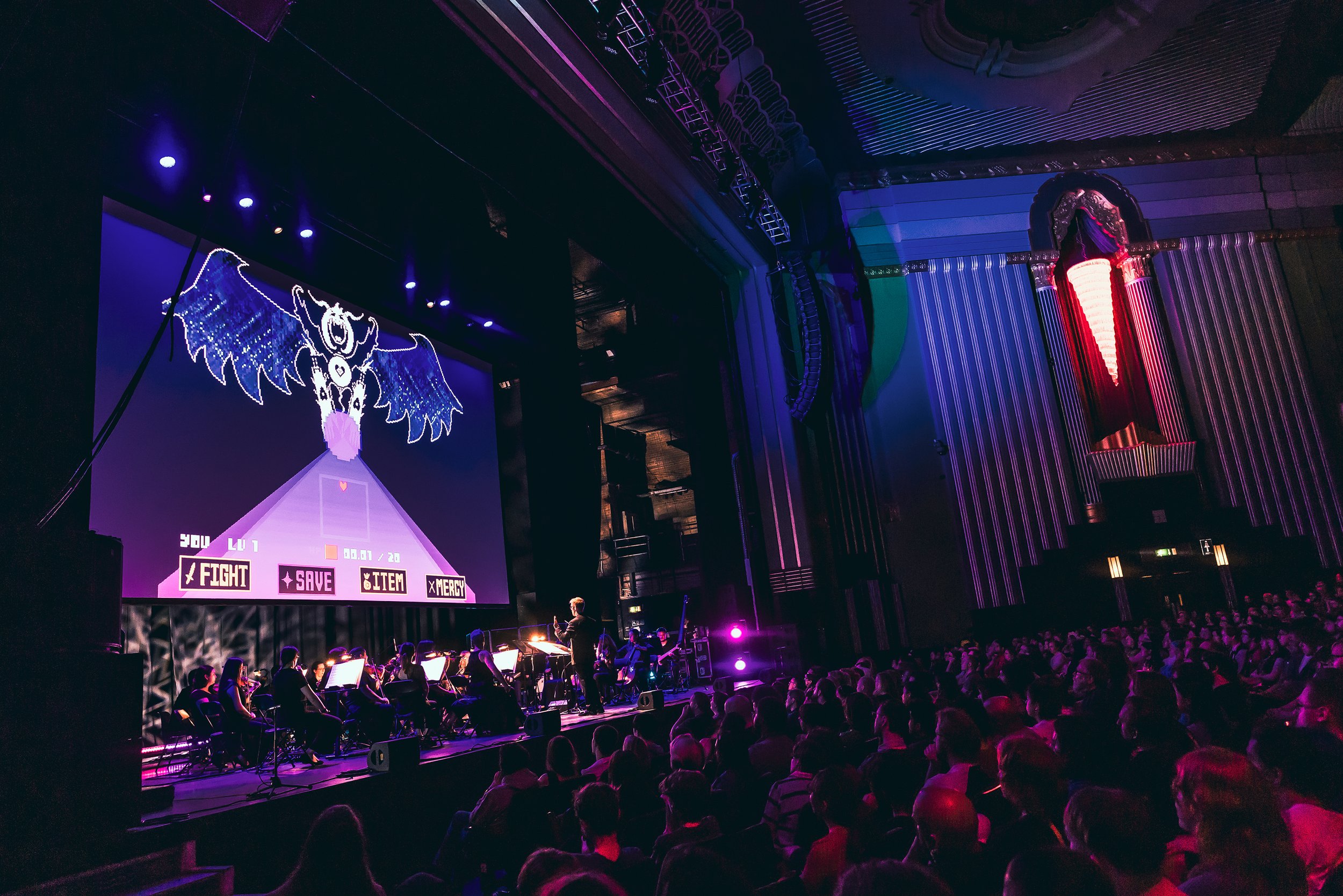 A theater or concert hall with a stage where a musical or theatrical performance is happening, featuring a large screen displaying a Undertale character or game interface, and an audience seated in front of the stage under colorful, dim lighting.