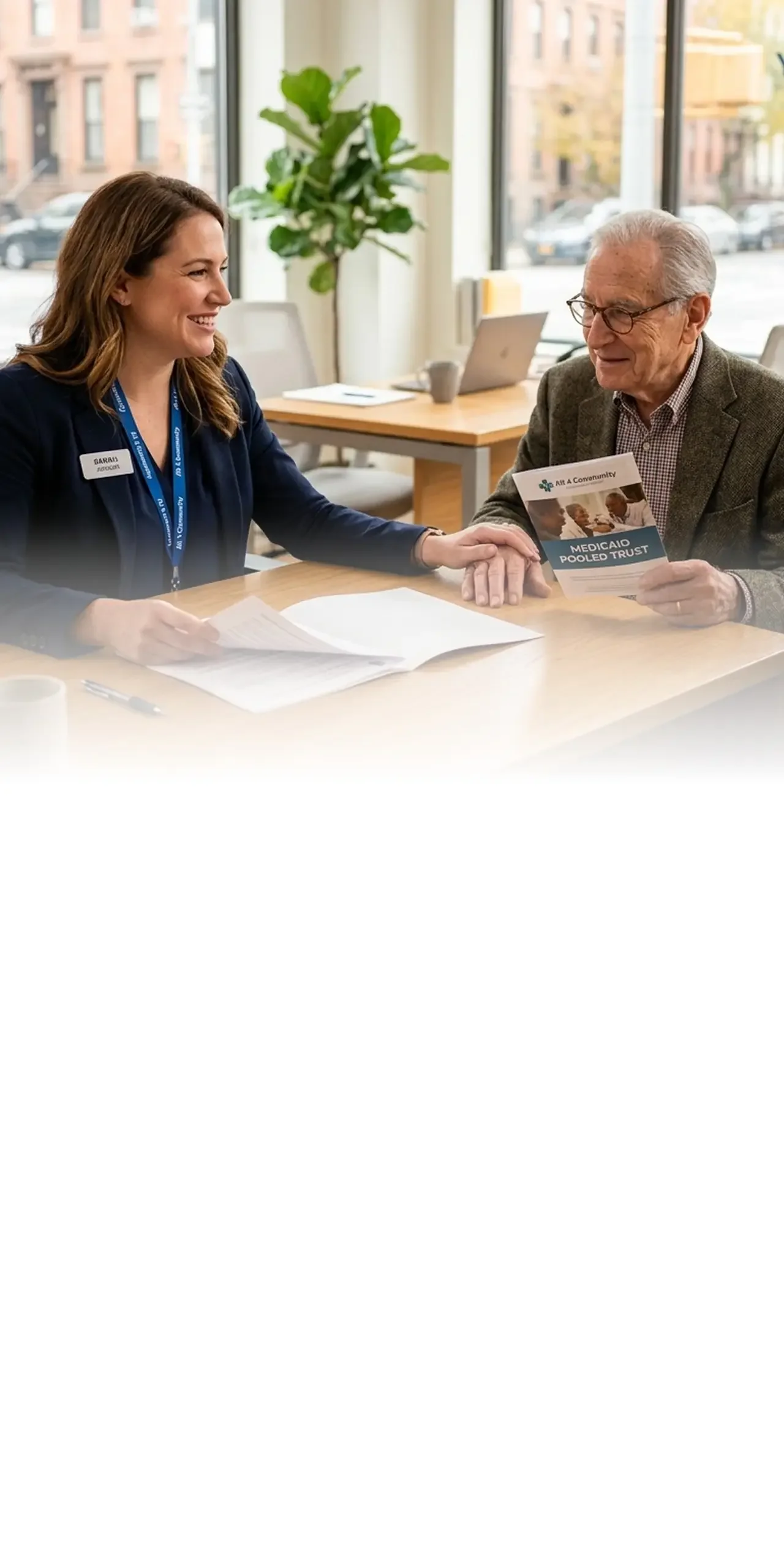Care coordinator sitting at a desk and smiling while assisting a senior man holding a Medicaid Pooled Trust brochure.