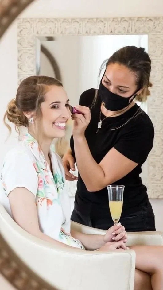 A woman in a floral robe smiling while getting her makeup done by a makeup artist wearing a black mask and shirt. The woman holds a glass of champagne.