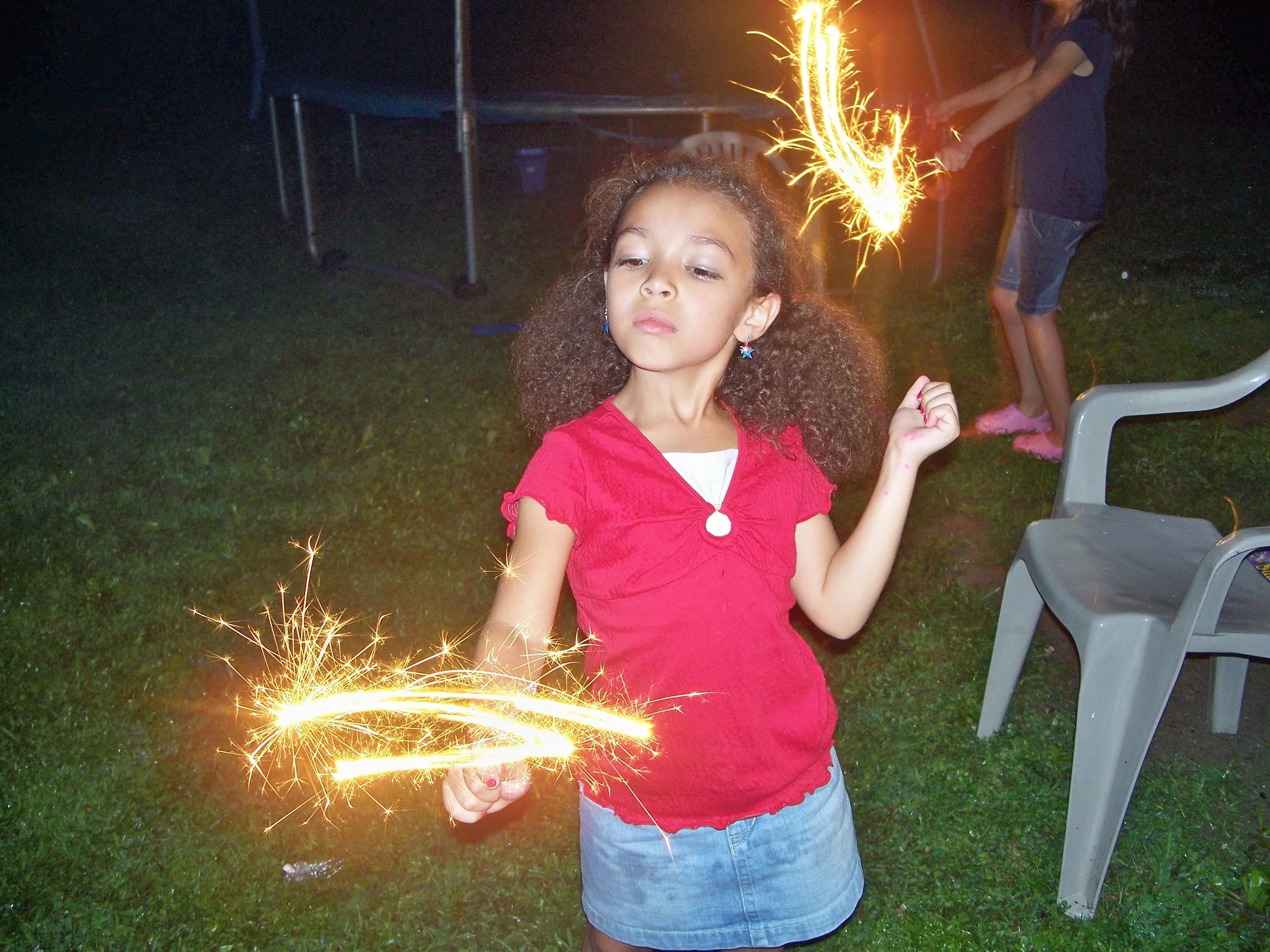 Young girl holding a lit sparkler at night, with another person in the background also holding a sparkler.