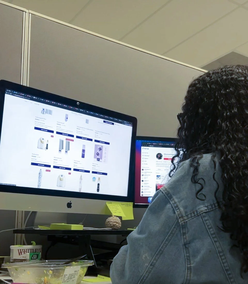 A person with curly dark hair wearing a denim jacket working at a desk with a computer monitor displaying an online store with household appliances and products.