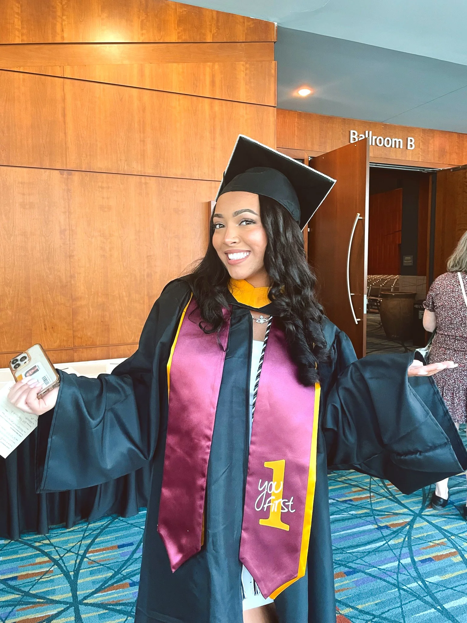 A young woman in graduation cap and gown celebrating her graduation in a large indoor space, holding a smartphone and a diploma, with a wooden wall and open door labeled "Ballroom B" in the background.