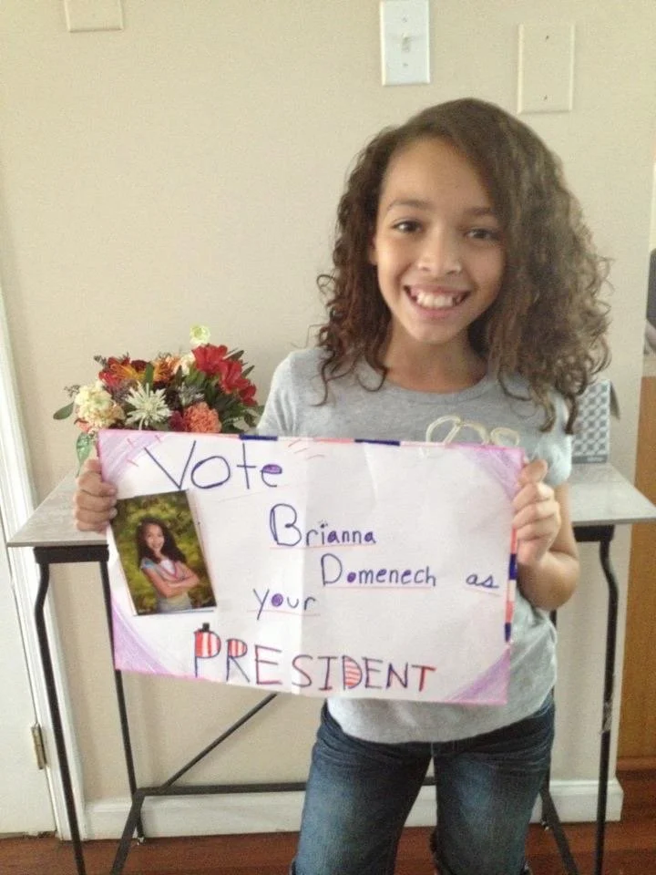 A young girl with curly hair is smiling and holding a handmade sign that says "Vote Brianna Domenech as your PRESIDENT" with a small photograph of herself attached. There is a flower arrangement on a table behind her.