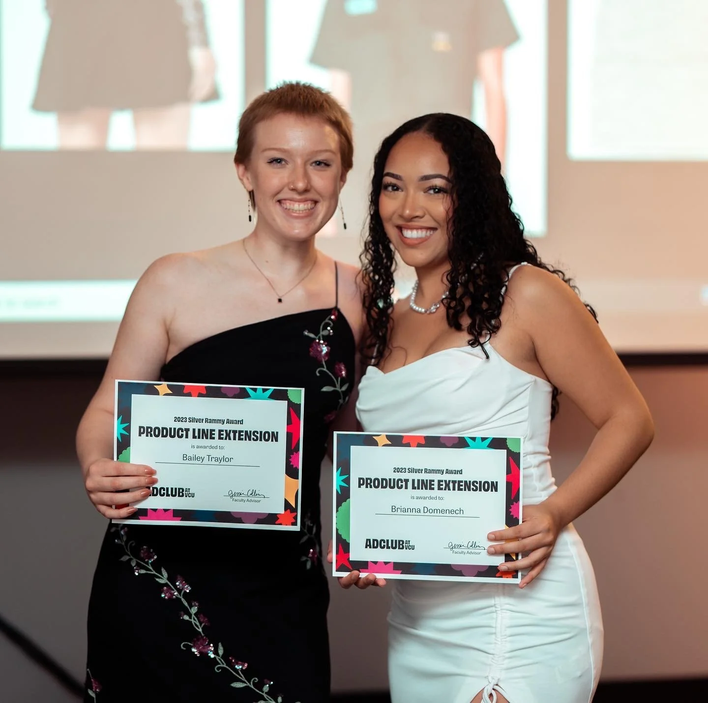 Two women smiling and holding certificates at an award event, with a projector screen in the background.
