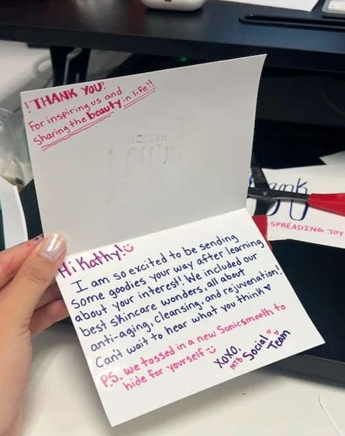 A handwritten thank you note on white paper, with pink and blue ink, expressing excitement about sending skincare goodies and including a PS about a new SonicSmile device, signed by an MTB Social Team member.