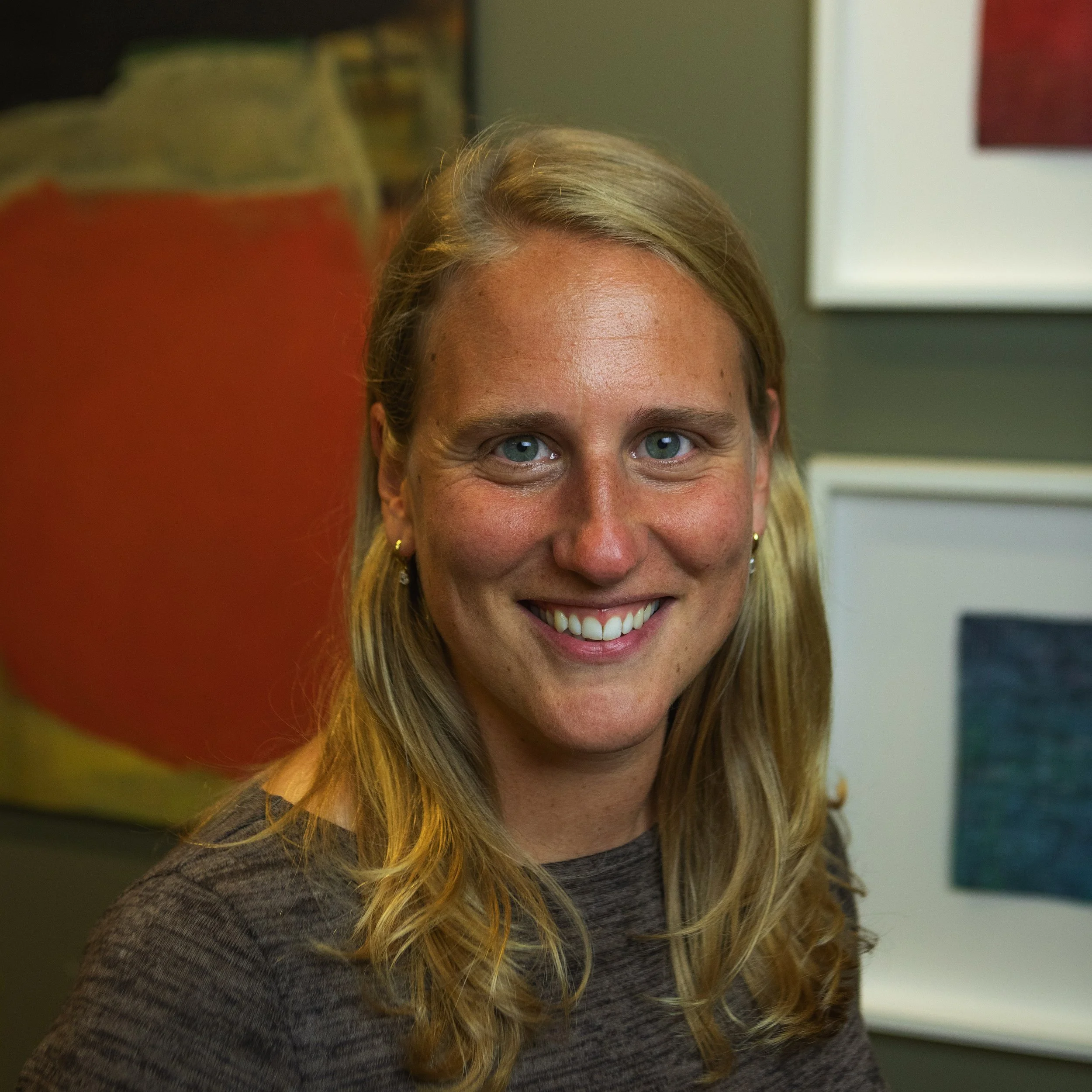 Close-up portrait of a smiling woman with long blonde hair, blue eyes, and wearing a gray top, standing indoors with artwork in the background.