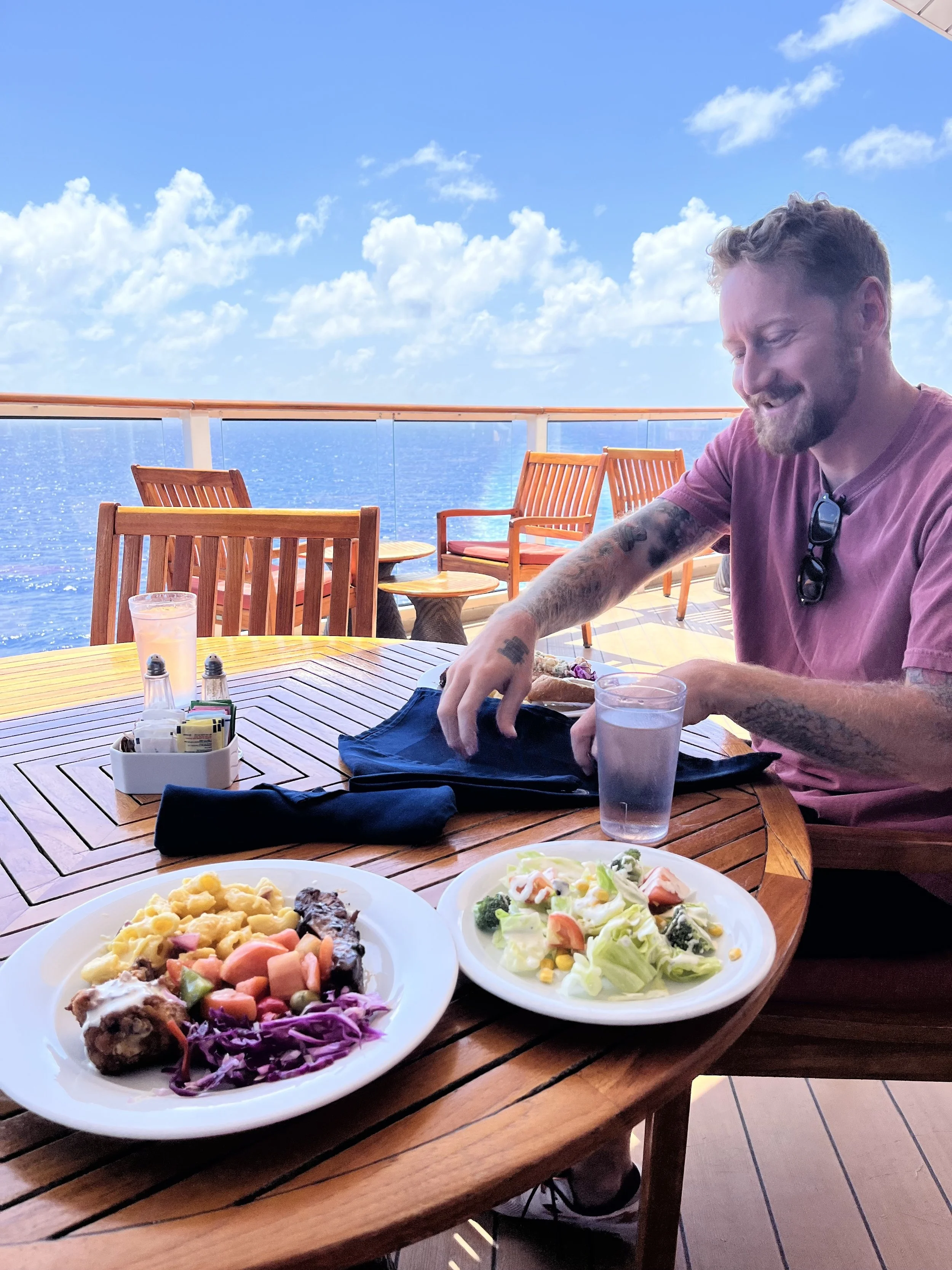 A man sitting at a round wooden table on a cruise ship deck during daytime, with two plates of food, a glass of water, and a black cloth on the table, overlooking the ocean with blue sky and clouds.
