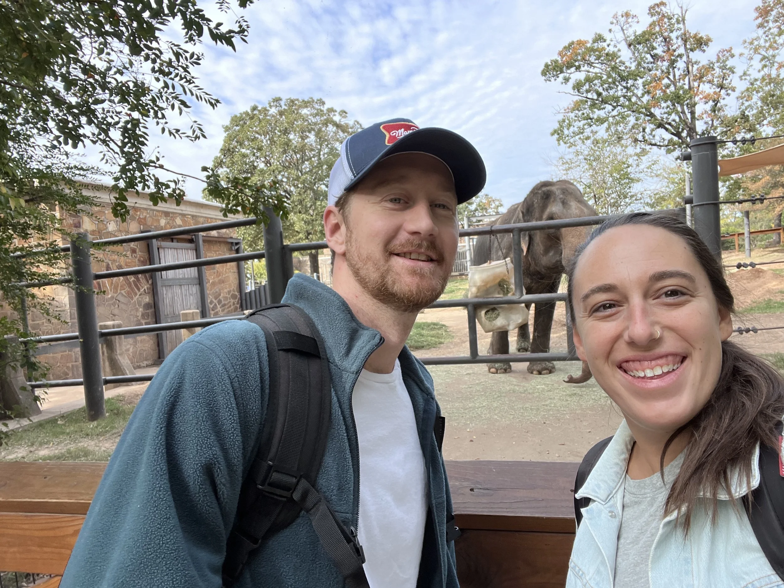 A smiling man and woman taking a selfie at a zoo with an elephant in the background behind a fence, trees, and a partly cloudy sky.