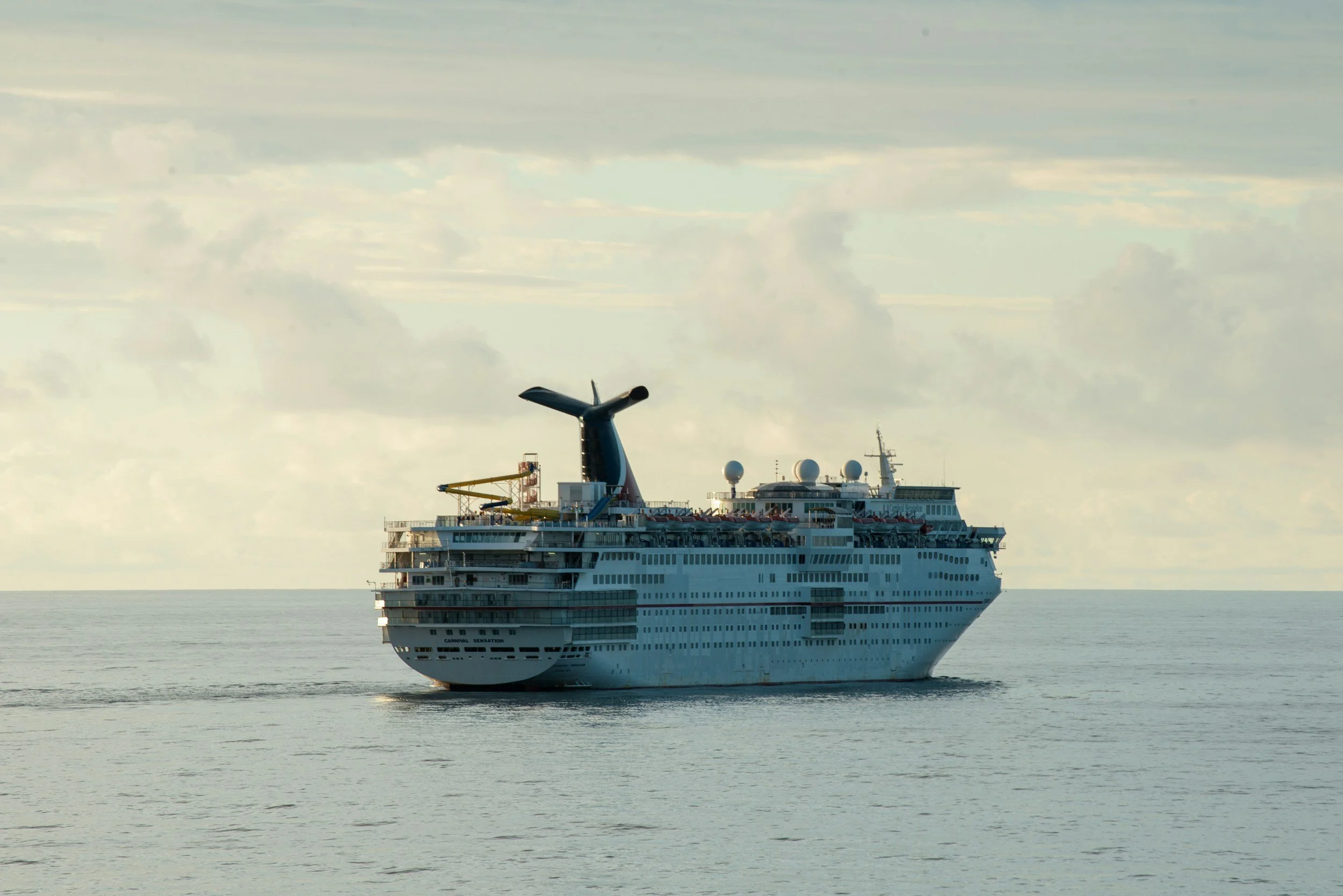 Large white cruise ship sailing on calm ocean with cloudy sky in background.
