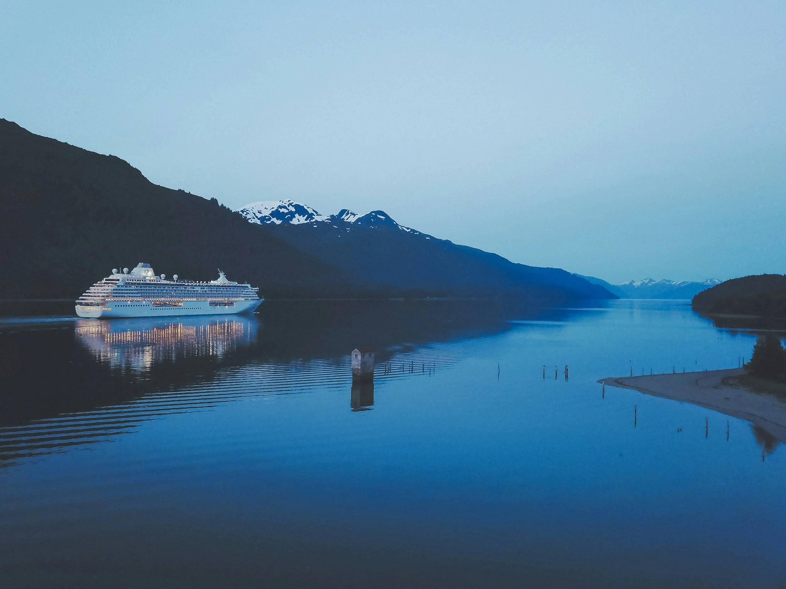 A large white cruise ship sailing on a calm blue fjord surrounded by mountains with snow-capped peaks at dusk.
