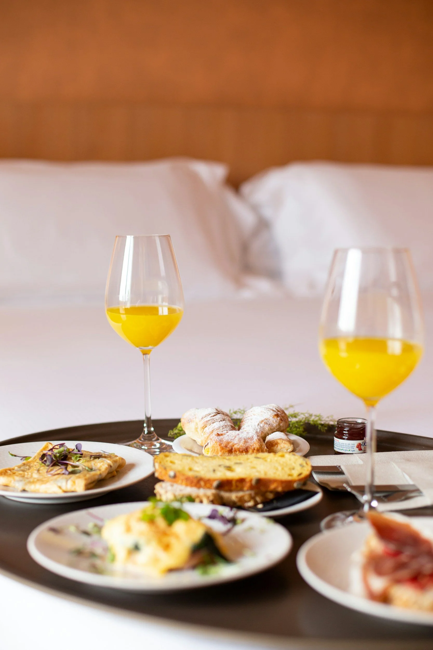 Breakfast in bed with two glasses of orange juice, pastries, bread, and breakfast dishes on a tray in front of a bed with white pillows.