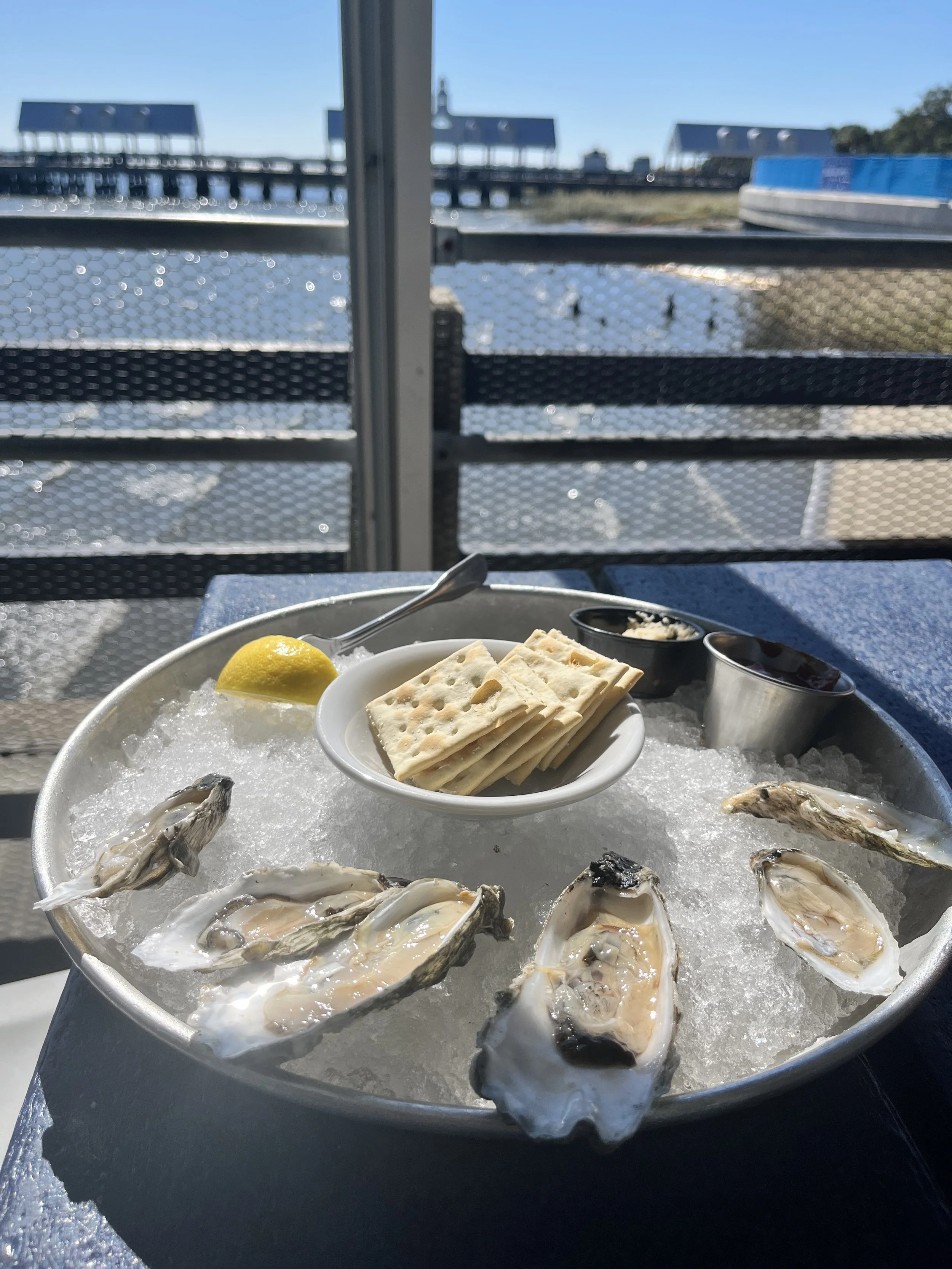 A plate of fresh oysters on ice with lemon wedge, crackers, and sauces at an outdoor seafood restaurant by the water.