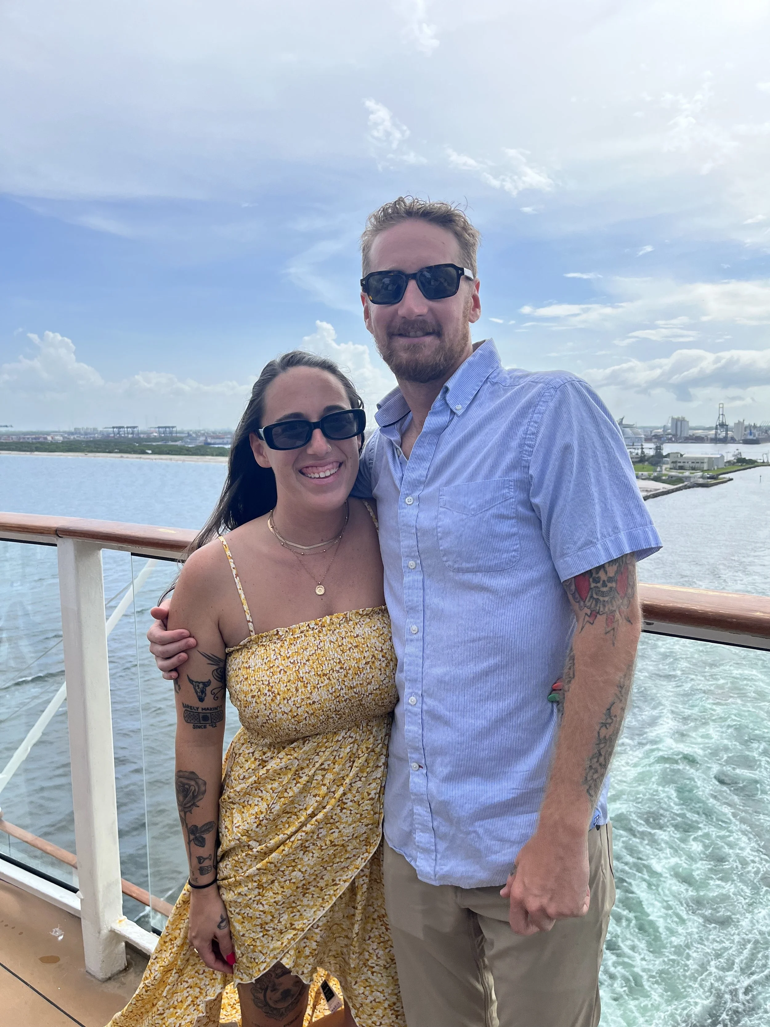 A smiling couple wearing sunglasses standing on a cruise ship deck with water, a distant shoreline, and a cloudy sky in the background.