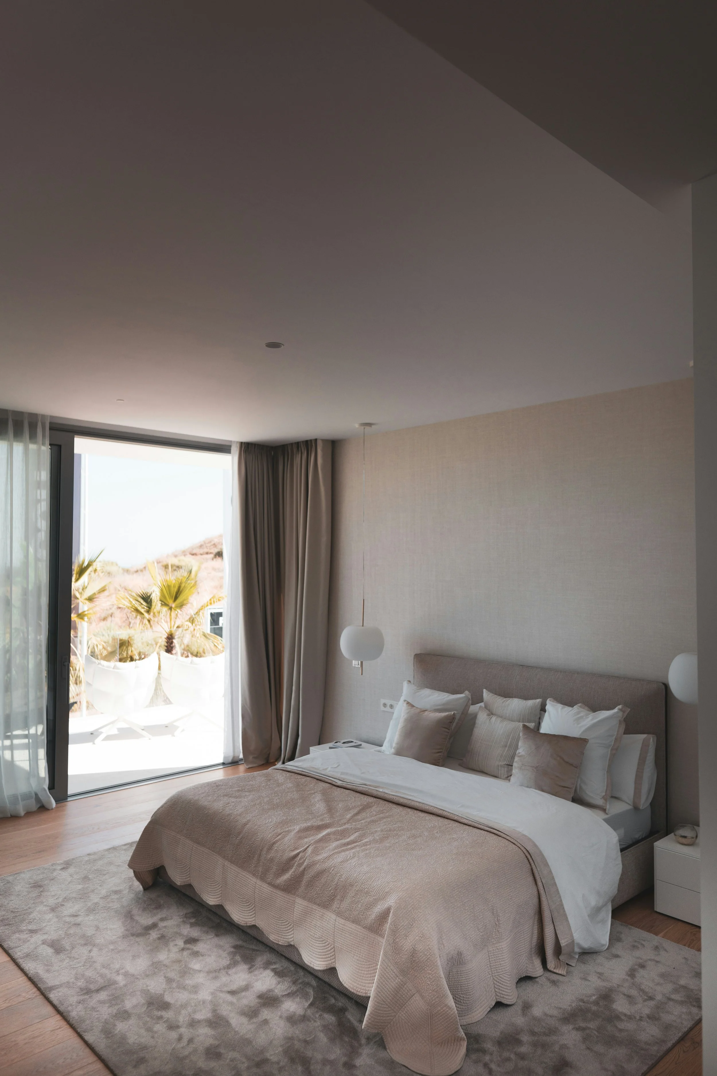 Hotel bedroom with king-sized bed, beige bedding, and multiple pillows, next to a large window with sliding glass door and curtains, overlooking outdoor palm trees.
