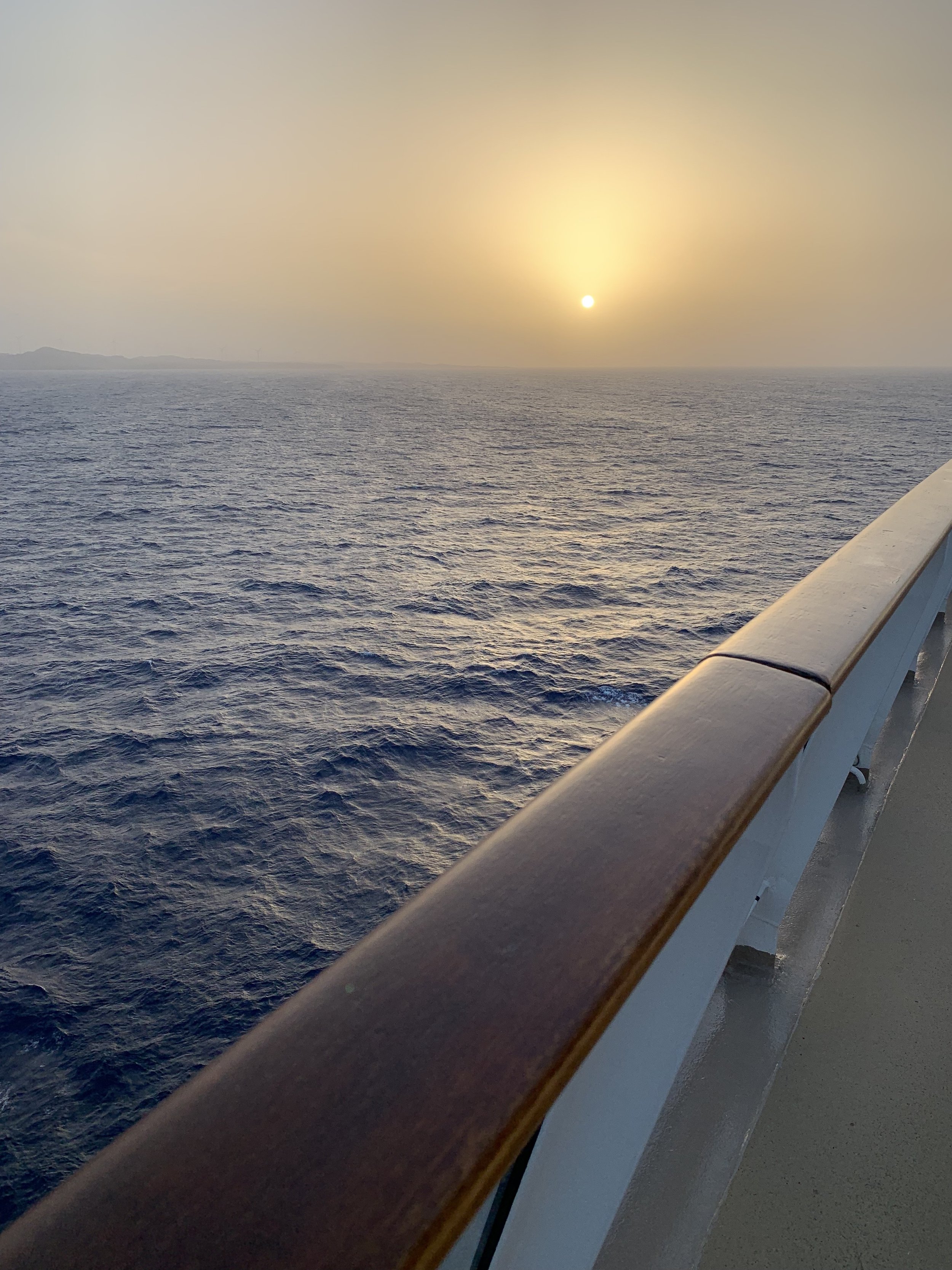 View of the ocean at sunset from a ship deck, with a wooden railing and distant land on the horizon.