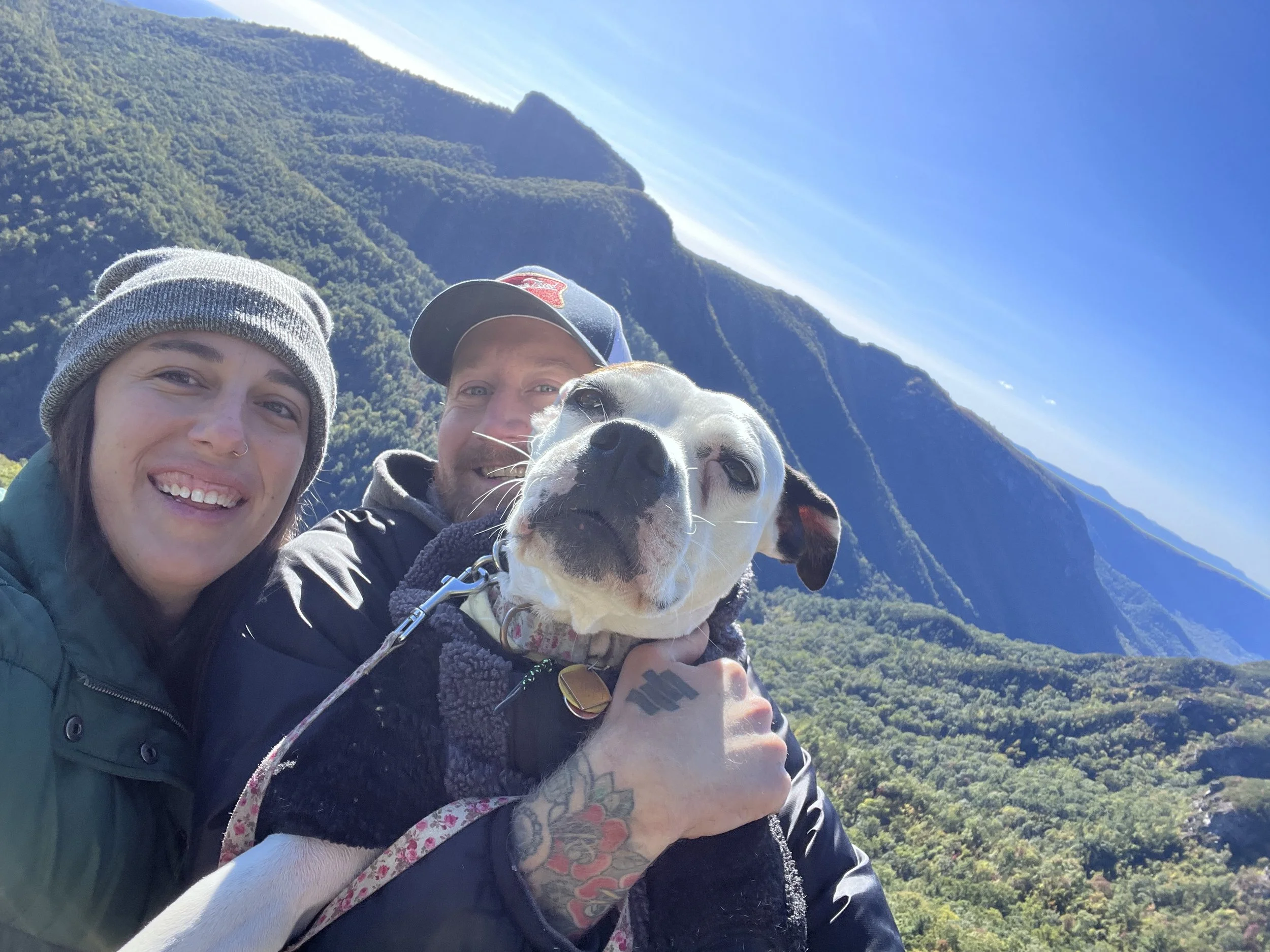 Two people smiling and a dog taking a selfie with a mountain landscape in the background.