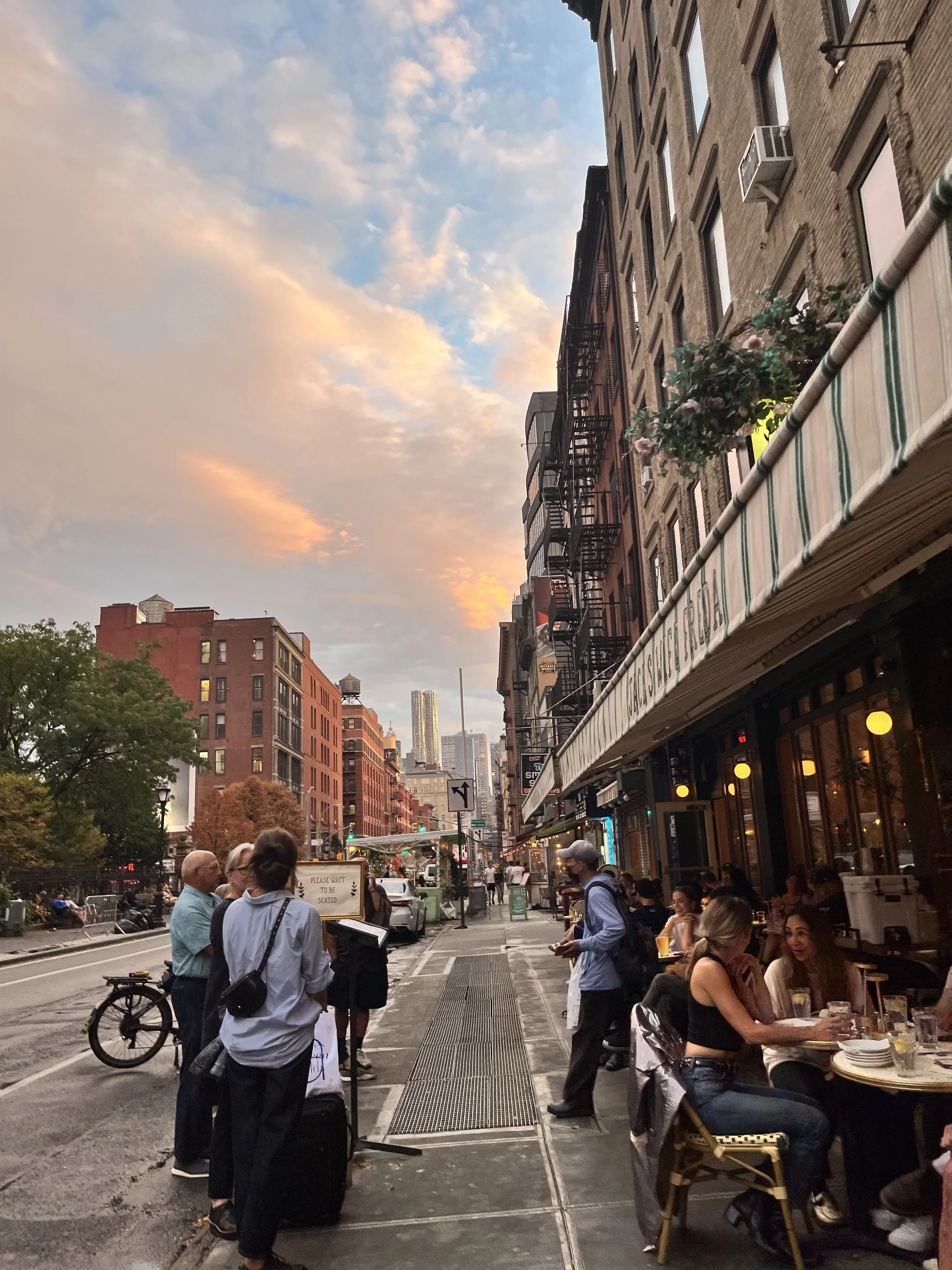 People dining and socializing outside a restaurant on a city sidewalk at sunset, with buildings and city skyline in the background.