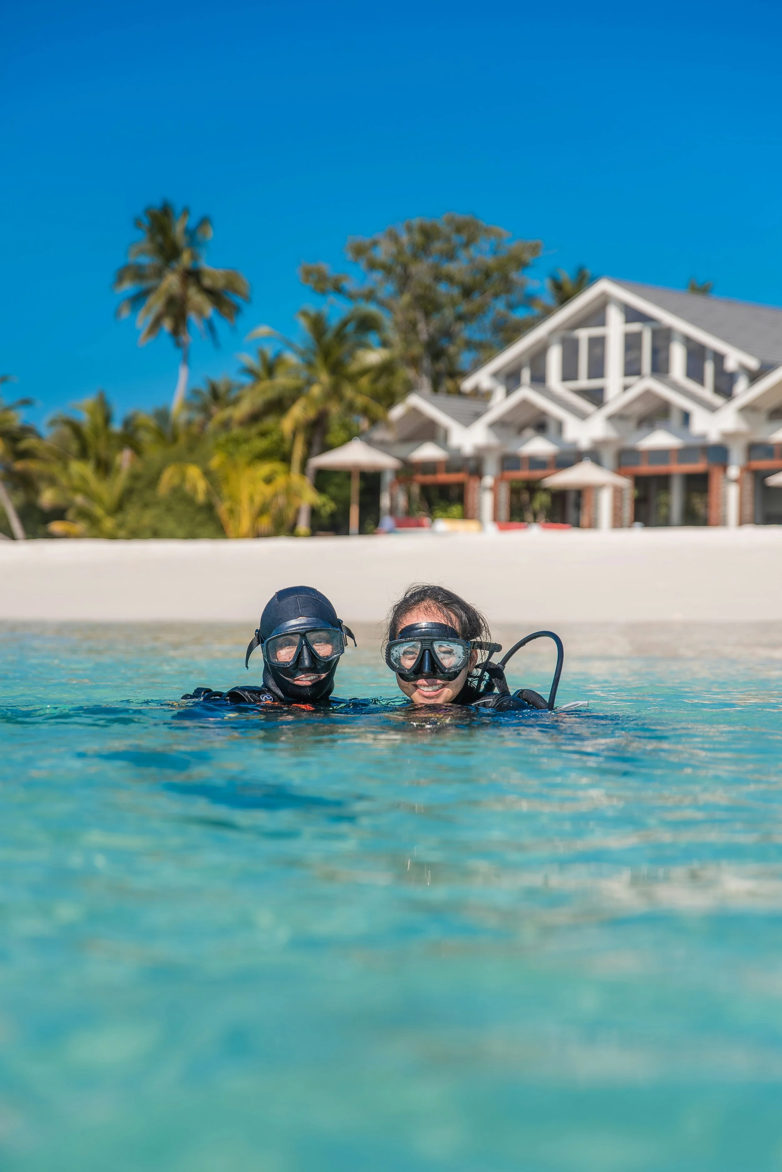 Two women in scuba diving gear smiling in shallow clear water with a tropical beach house and palm trees in the background.