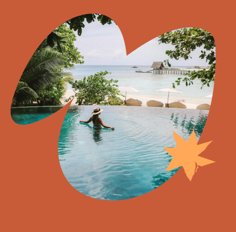 Woman with a sun hat relaxing in a swimming pool overlooking a beach with umbrellas and a pier, surrounded by lush trees, under a partly cloudy sky.