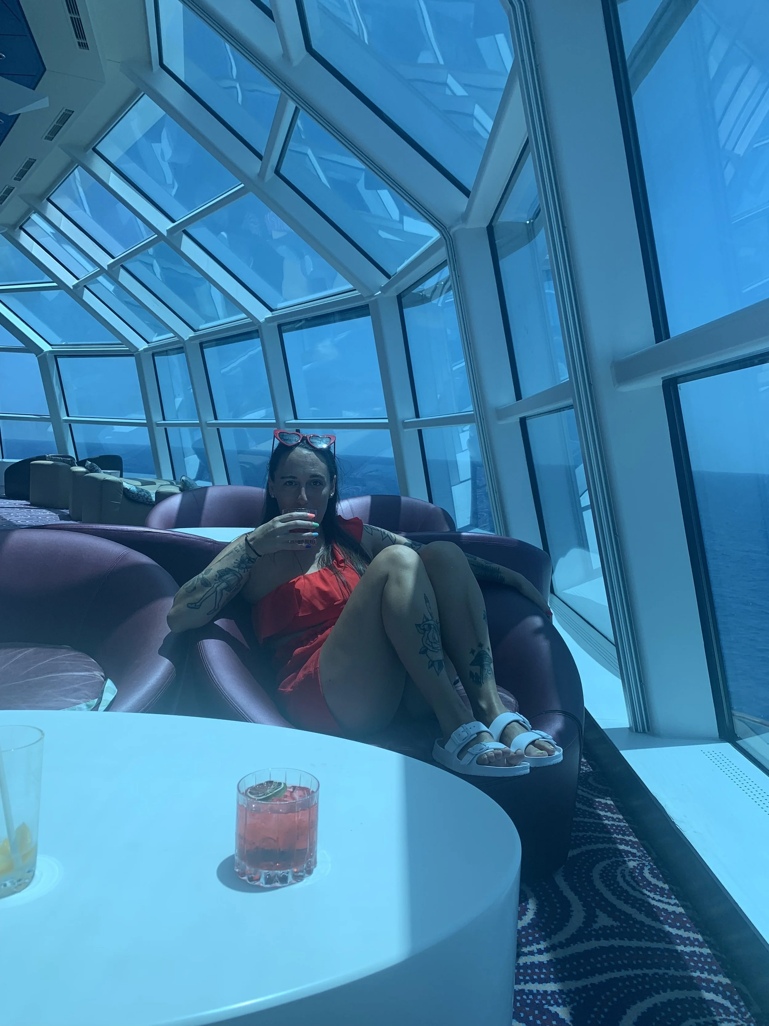 Woman in a red dress and white sandals sitting on a lounge chair inside a glass-walled room, drinking a beverage, with drinks on a white table in the foreground.
