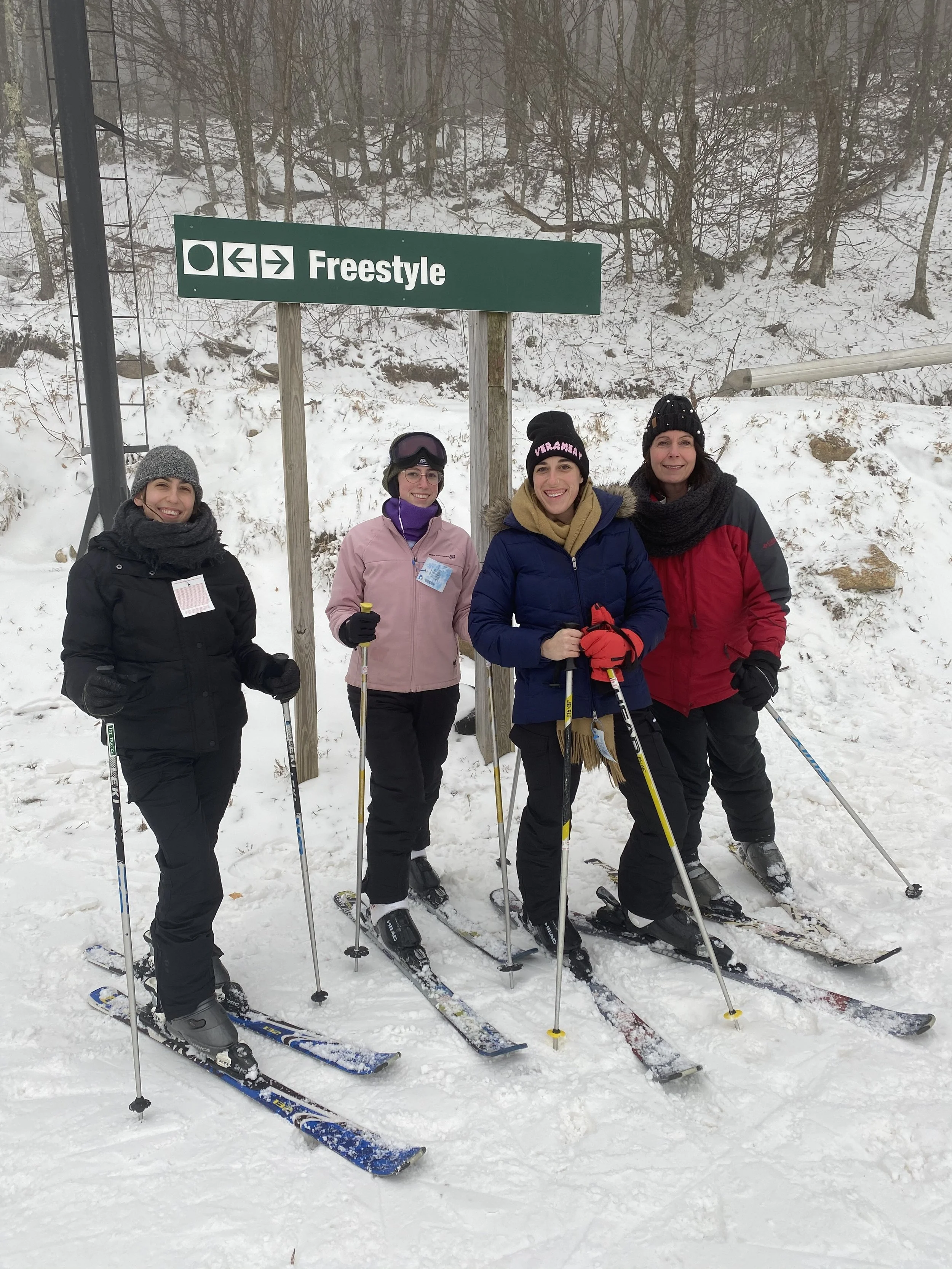Four women in winter clothing standing on skis in the snow, smiling, with a green sign that says 'Freestyle' and arrow directions behind them.