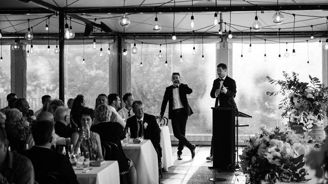 A black and white photo of a formal event or wedding reception with guests seated at tables. Two men in tuxedos are standing at a podium; one is speaking while the other is listening. Decorative flowers and hanging string lights are visible.