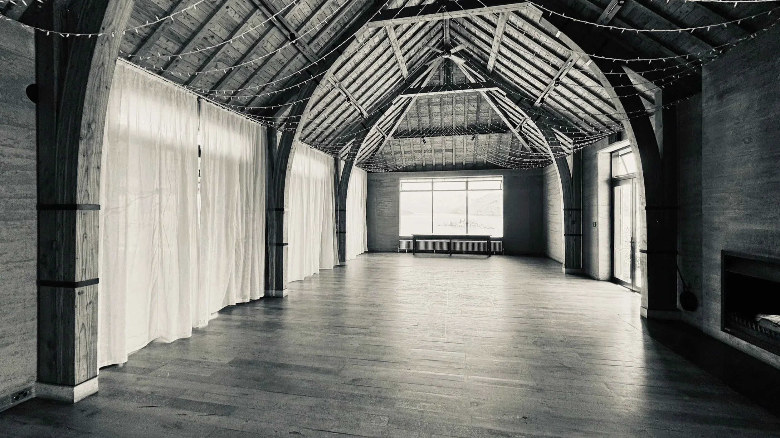 Empty rustic event hall with wooden floors, exposed wooden beams, and string lights hanging from the ceiling. There are large windows and white curtains along one side.