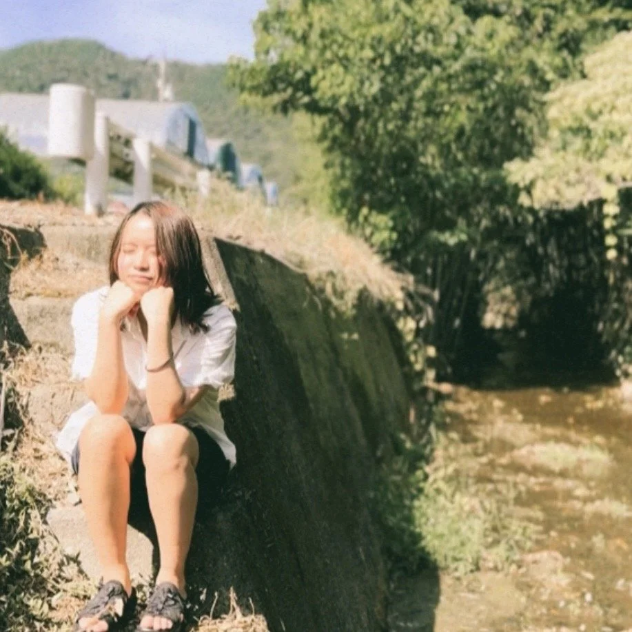 A girl sitting on a concrete ledge next to a small creek or stream, surrounded by greenery and trees, with a clear sky in the background.