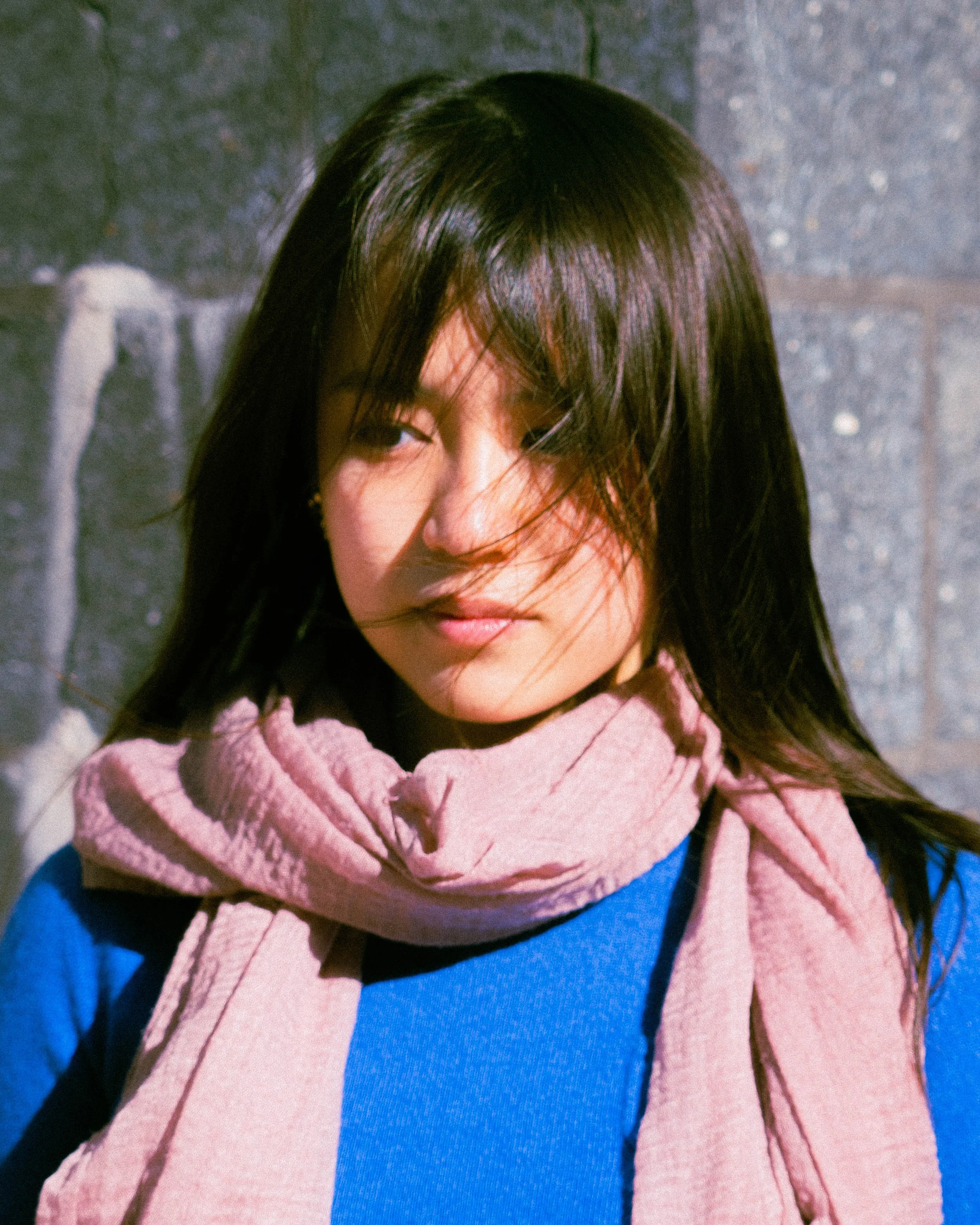 A young woman with long dark hair partially covering her face, wearing a pink scarf and blue top, standing outdoors in front of a wall with graffiti.