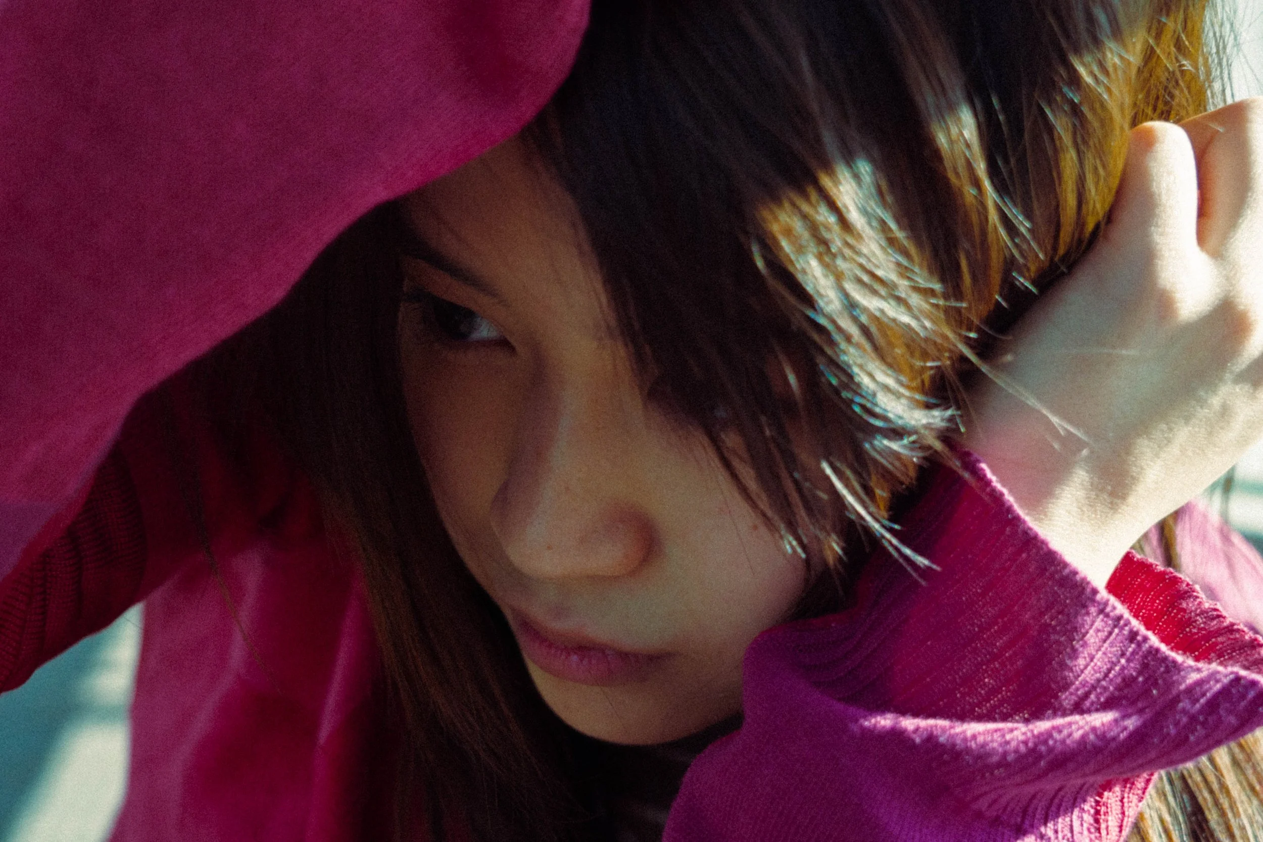 A close-up of a young woman with brown hair partially covered by a pink hoodie, resting her head on her arm and looking to the side, in natural lighting.