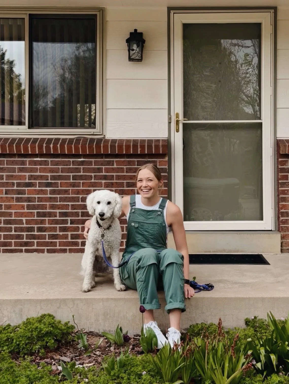 A young woman sitting on a concrete porch with a white poodle dog, both smiling. The woman wears green overalls, white sneakers, and a white tank top. The porch is in front of a house with a brick lower wall, white upper wall, and glass door. There are flowers and green plants in front of the porch.