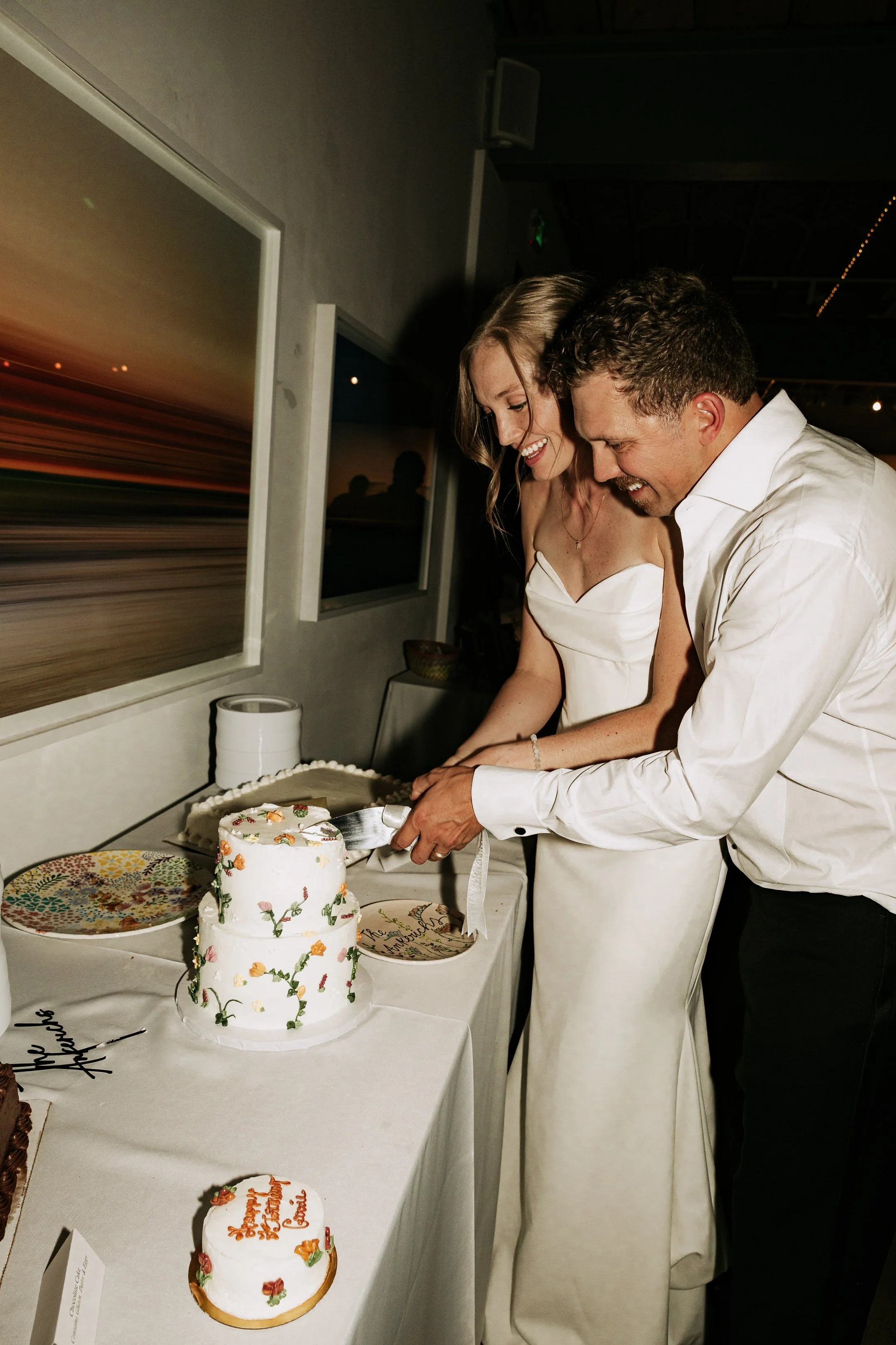 A newlywed couple in wedding attire cuts their wedding cake at a celebration.