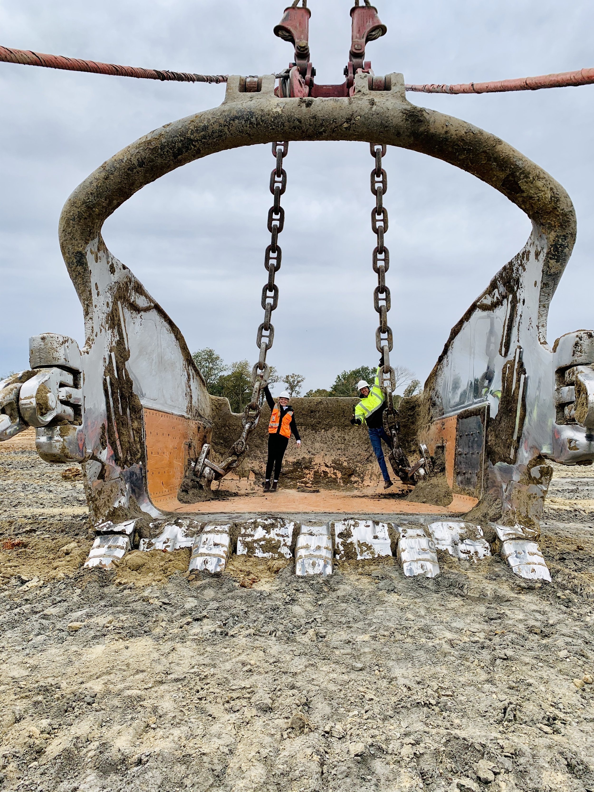 Close-up of a large construction excavator bucket with two workers in safety gear and helmets inside, visible through the opening, on a construction site with dirt and an overcast sky.
