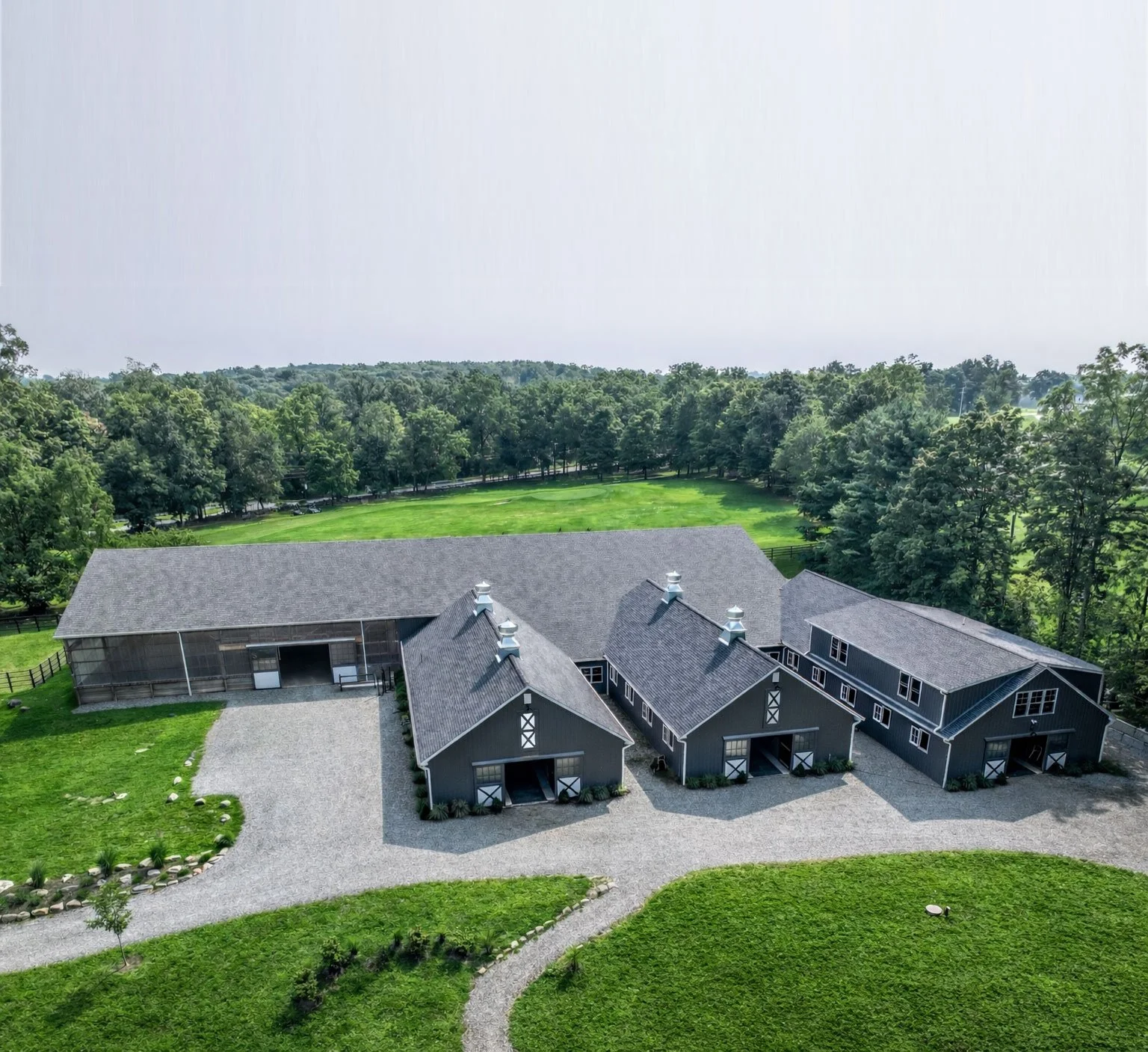 A large modern gray barn with multiple sections and a gravel driveway in front, surrounded by green lawn and trees, with open fields and a woods background.