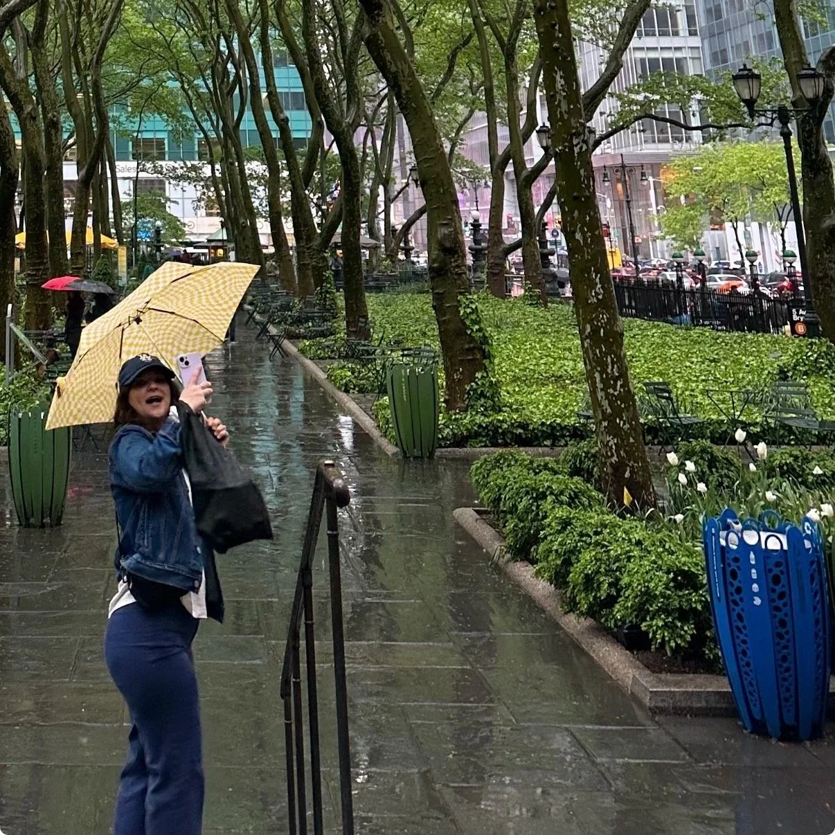 Woman with a yellow checkered umbrella talking or singing on a rainy city street, surrounded by trees, benches, and plants, with buildings and cars in the background.